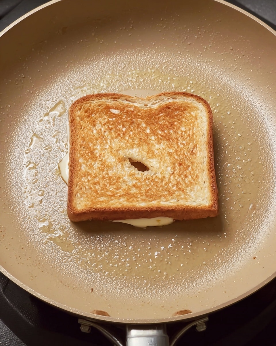 A single sandwich with two light brown toasted bread slices is placed flat in the center of a beige frying pan. The sandwich's top slice shows a slight golden texture with a small hole near the middle, and melted white cheese is peeking out thinly from the edges underneath. The frying pan surface has a shiny layer of butter or oil spread evenly around the sandwich, creating a subtle glistening effect. The pan is positioned on a stove, with the pan's handle partially visible at the bottom. The overall look is warm and ready for grilling. photo taken with an iphone --ar 4:5 --v 7