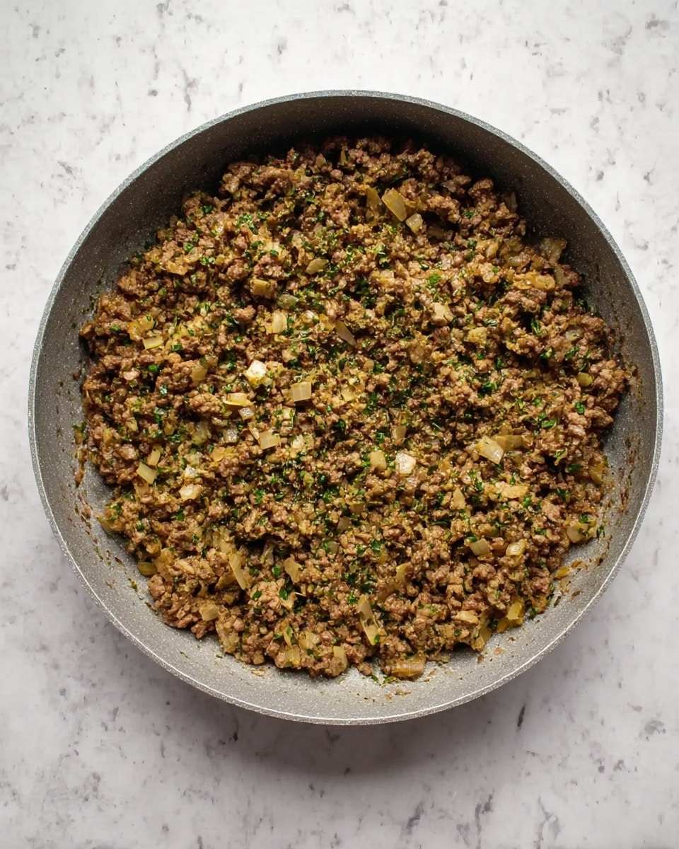 A round gray pan filled with cooked ground meat mixed with small pieces of cooked onion and green herbs, evenly spread across the pan. The texture looks crumbly and moist with a mix of brown, pale yellow, and green colors. The pan is placed on a white marbled surface. photo taken with an iphone --ar 4:5 --v 7