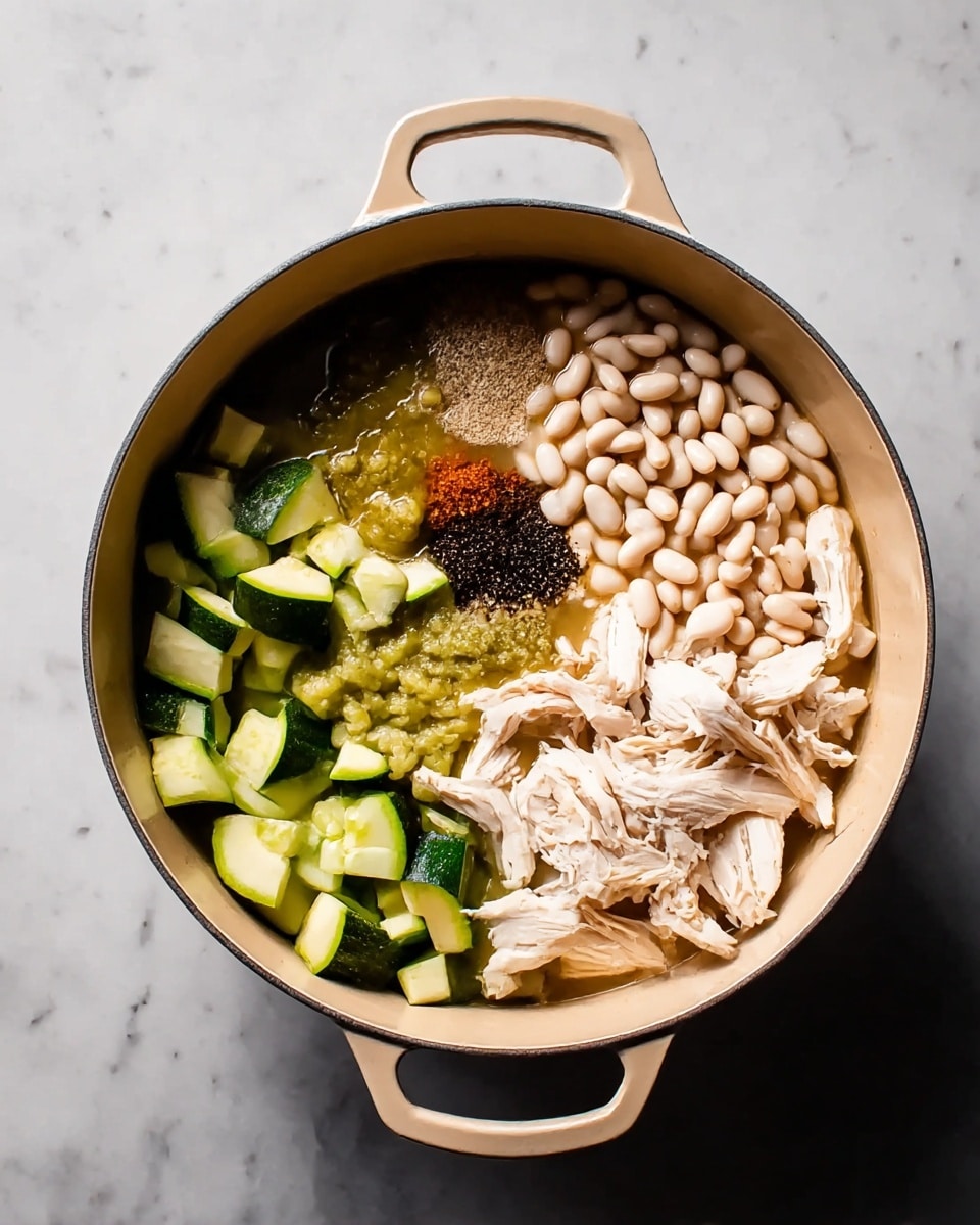 A large beige pot filled with layers of raw ingredients arranged side by side is shown from above on a white marbled surface. Starting from the bottom right, there are shredded pale pieces of chicken meat, next to it toward the top right, a pile of small white beans partly submerged in a light broth. Moving left from the beans, there is a dark brown spice powder and some black pepper next to it. The left side of the pot shows bright green cubed vegetables below a mix of sliced pale green zucchini and chunky green sauce. The pot has two handles on each side. photo taken with an iphone --ar 4:5 --v 7