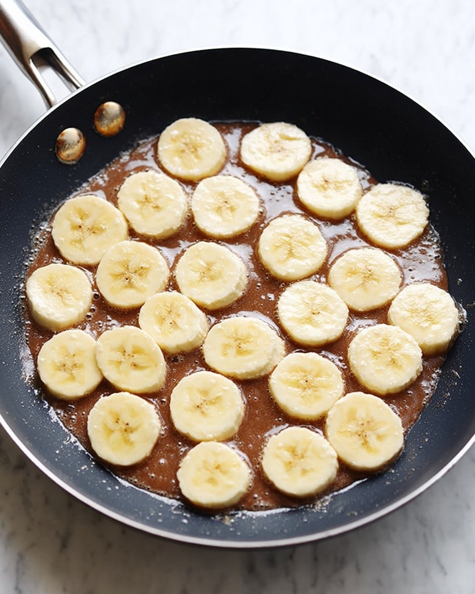 A close-up top view of a dark non-stick frying pan filled with a thin layer of brown batter spread evenly on the bottom. On top, there are 20 round slices of banana, light yellow in color with a soft texture, scattered in a loose pattern across the batter. The pan's metal rim and rivets are visible on the left side. The background is a white marbled surface. photo taken with an iphone --ar 4:5 --v 7