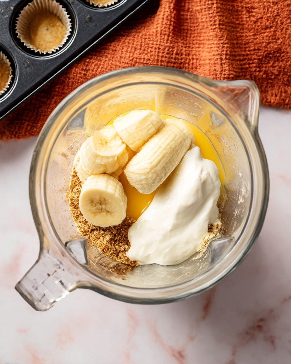 Inside a clear glass blender container, a mixture is arranged with three layers of light yellow banana pieces positioned centrally on top. To the right of the bananas, there is a dollop of thick white yogurt with a smooth, creamy texture. Underneath and partially visible, there is a layer of golden yellow liquid, likely egg yolk, resting in the back center. Surrounding the bananas and yogurt, on the bottom layer, is a mix of brown powder and granules, giving a rough texture that contrasts with the smoothness above. The blender sits on a white marbled surface, with a textured burnt orange cloth nearby and a corner of a black muffin tray in the upper left background. photo taken with an iphone --ar 4:5 --v 7