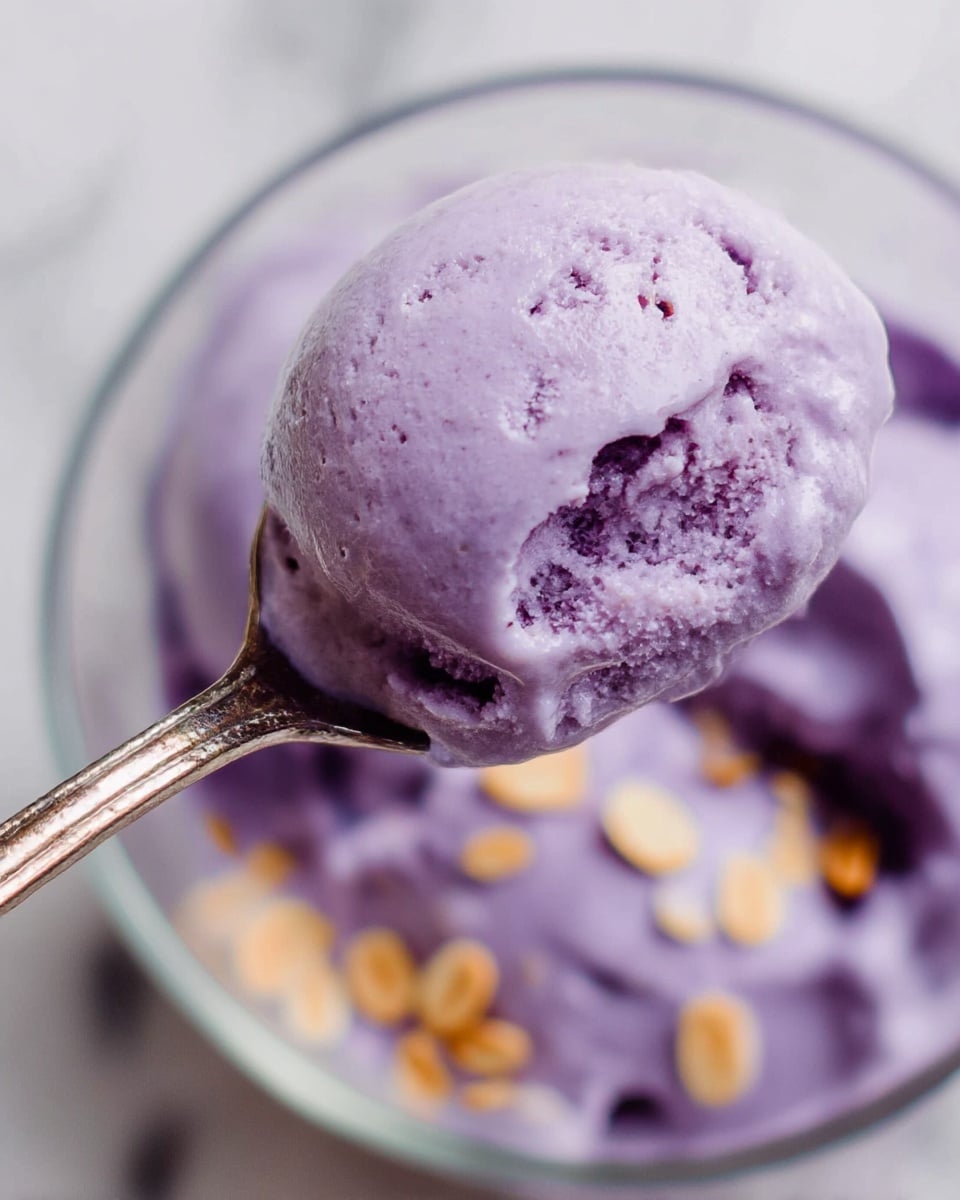 A close-up view of creamy purple ice cream being held by a spoon above a clear bowl. The ice cream looks smooth with a few small chunks inside, and some small light brown flakes are sprinkled on top and visible in the bowl below. The clear bowl sits on a white marbled surface, and the focus is on the spoon with the ice cream, showing its thick texture. Photo taken with an iphone --ar 4:5 --v 7