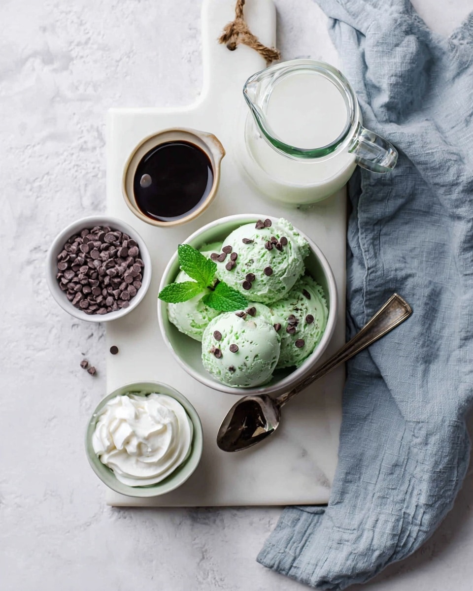 The image shows a white cutting board on a white marbled surface holding a white cup filled with three scoops of light green mint chocolate chip ice cream garnished with small chocolate chips and two fresh green mint leaves on top. Next to the cup is a silver spoon filled with dark chocolate syrup resting on the board. To the left of the cup, there is a small white bowl with dark chocolate chips. Above the cutting board, there is a clear glass pitcher filled with milk, and to the right of the pitcher is a small white bowl with a swirl of whipped cream. A light blue fabric napkin is draped casually over the white marbled surface on the right side. photo taken with an iphone --ar 4:5 --v 7