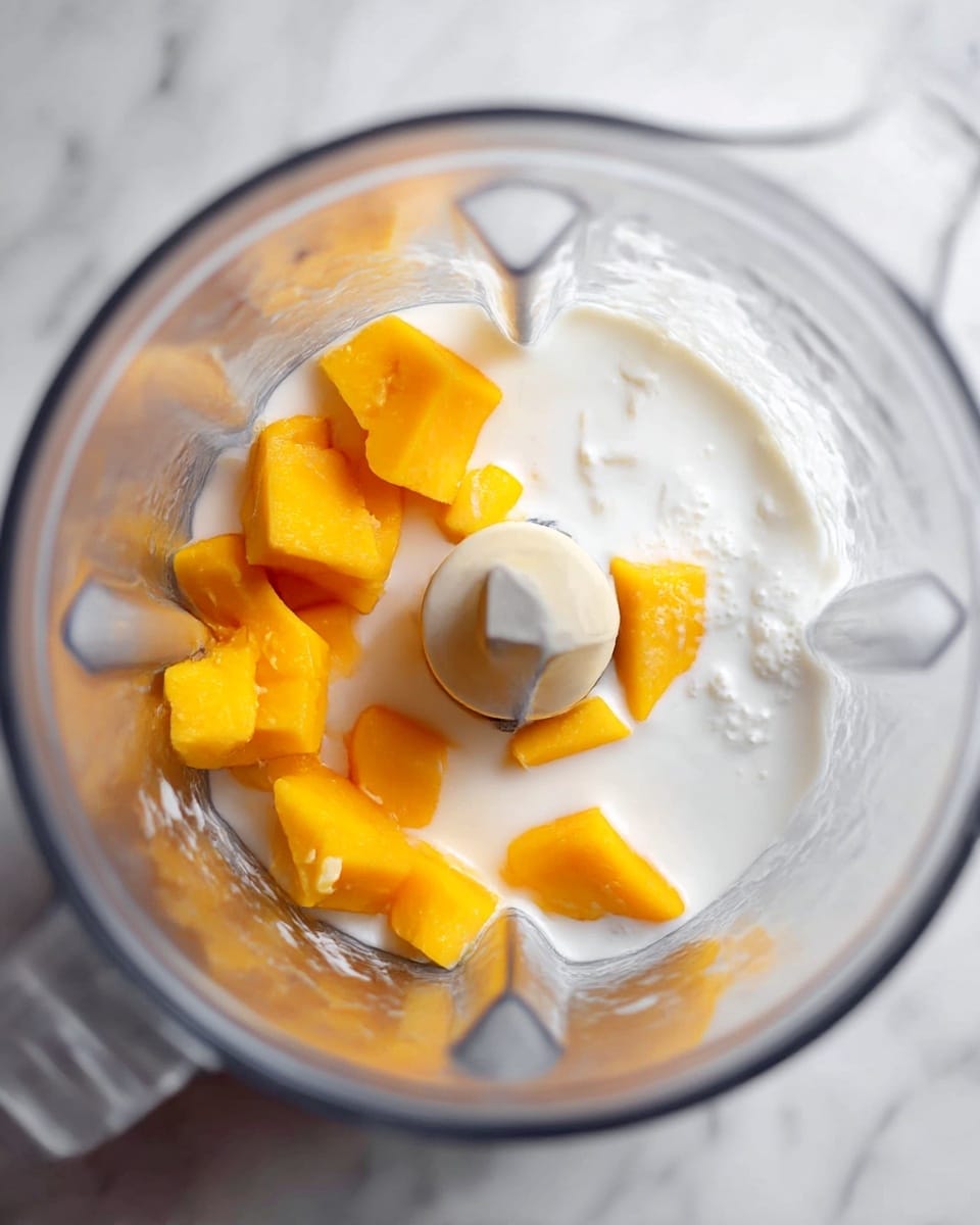 A top view of a clear blender jar showing chunks of bright orange mango sitting on a layer of white coconut milk inside. The mango pieces are uneven in size and clustered on one side, while the liquid coconut milk spreads around the base and partially covers the mango. The sharp blender blade sits centered at the bottom in between the mango and milk layers. The background is a white marbled texture. photo taken with an iphone --ar 4:5 --v 7