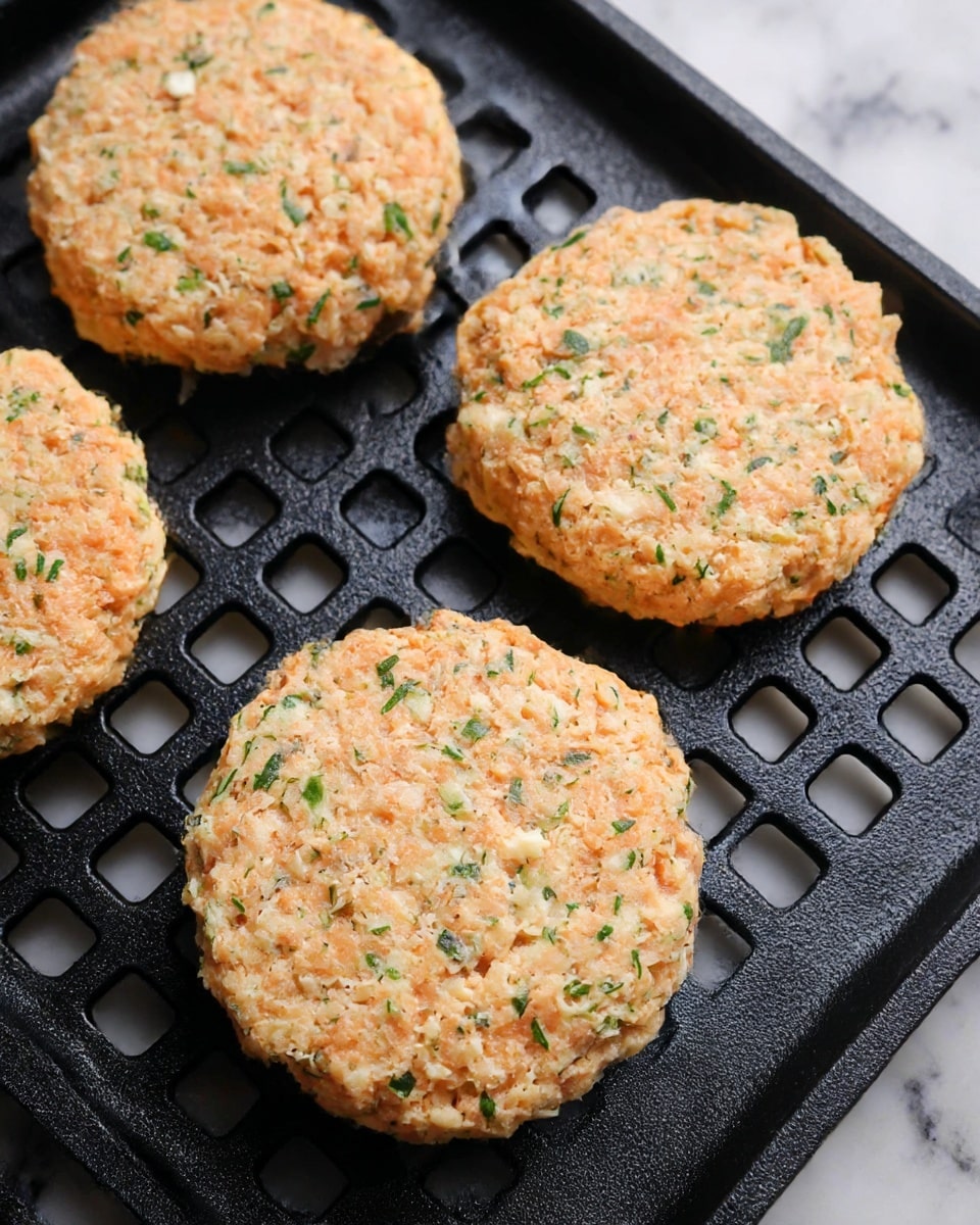 The image shows four round salmon patties with a rough texture, each mixed with small bits of green herbs and light seasoning, resting on a black grill pan with rectangular holes, placed on a white marbled surface. The patties have a pale orange color with specks of green herbs evenly spread throughout, and their edges appear slightly uneven. The grill pan has a matte finish and grid pattern, contrasting with the soft texture of the patties. photo taken with an iphone --ar 4:5 --v 7