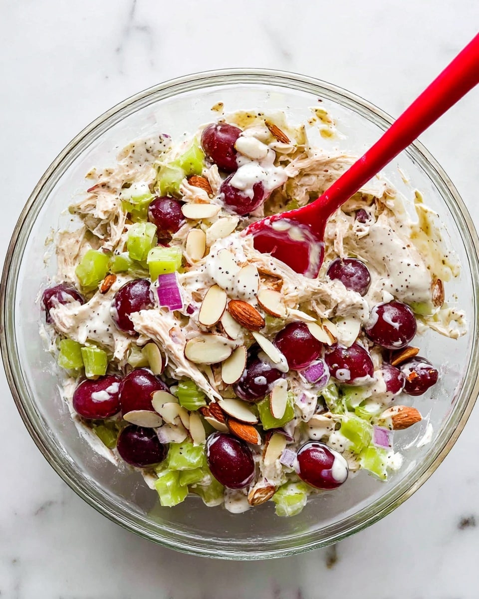A clear glass bowl filled with a colorful salad sits on a white marbled surface. Inside, there are layers of light beige shredded chicken, dark red halved grapes, light green diced celery, and small pieces of purple onion. Scattered over the top are thin, off-white almond slices and dollops of creamy white dressing with black pepper specks. A red spoon sticks out from the inside of the bowl, mixing the ingredients together. Photo taken with an iphone --ar 4:5 --v 7