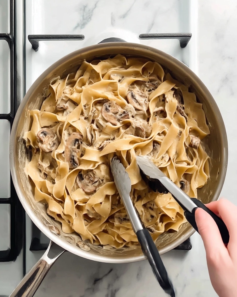 A silver cooking pan filled with wide, flat beige pasta noodles mixed with sliced brown mushrooms coated in a light creamy sauce. Woman's hand is holding black and silver tongs stirring the pasta. The pan is resting on a stove with a white marbled surface around it. photo taken with an iphone --ar 4:5 --v 7