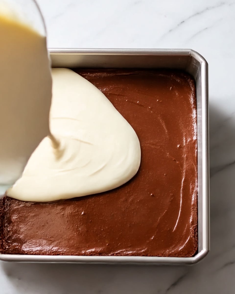 The image shows a square metal pan filled with a dark brown smooth chocolate layer, which covers the whole pan evenly. On top, a light cream layer is being poured over the chocolate layer, with the cream liquid spreading out smoothly and softly on one side of the pan. The background is a white marble surface. photo taken with an iphone --ar 4:5 --v 7