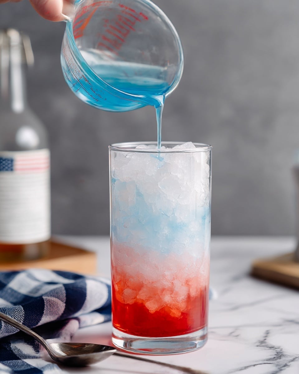 A clear glass filled with three layers of crushed ice in different colors: the bottom layer is bright red, the middle layer is white with slight pink shading, and the top layer is a pale blue. A measuring cup is held above the glass, pouring a blue liquid over a silver spoon to gently drip onto the ice. The background includes a blurry bottle with a white label and a checkered cloth in the foreground on a white marbled surface. Photo taken with an iphone --ar 4:5 --v 7