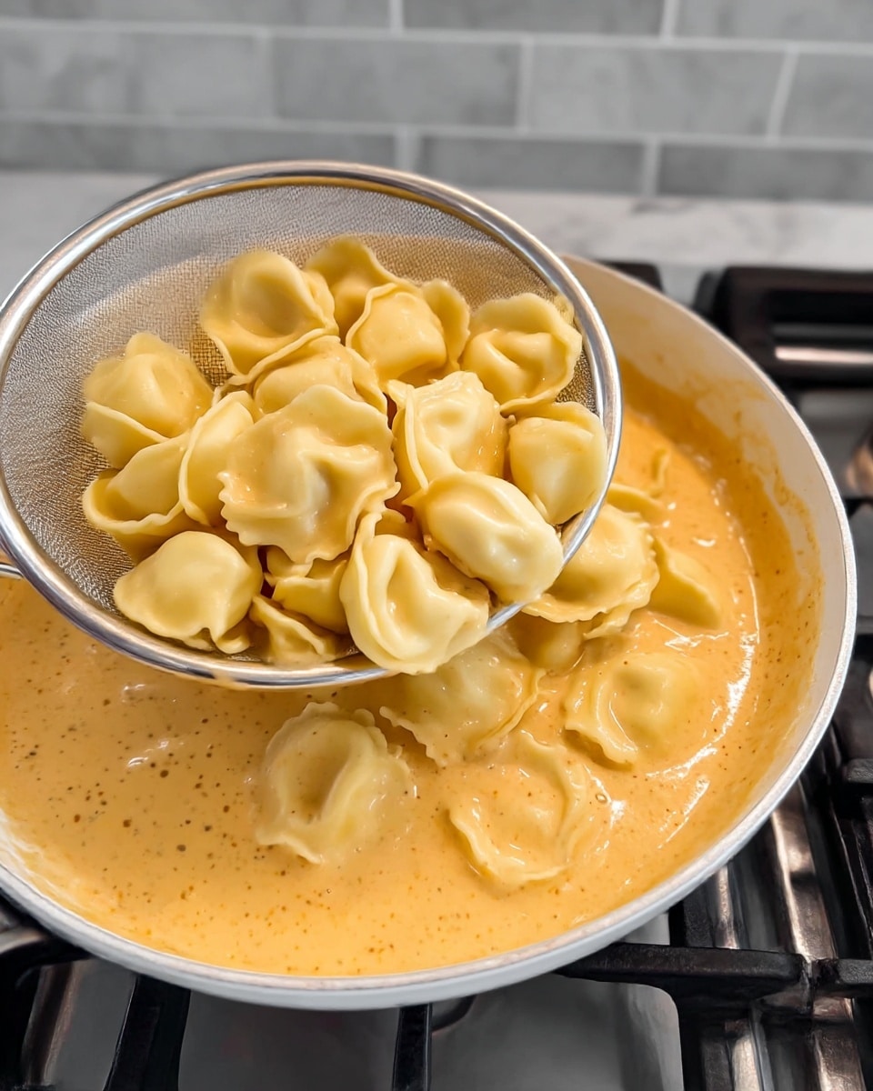 A close-up of cooked tortellini pasta with a smooth yellow color and soft texture, being held in a silver metal strainer near the top left corner. The pasta is about 20 pieces with a round shape and folded edges. Underneath, there is a creamy orange sauce with a thick and slightly speckled texture filling a white pan. The pan is on a gas stove with black grates, and the background is a white marbled surface with gray tiles. photo taken with an iphone --ar 4:5 --v 7