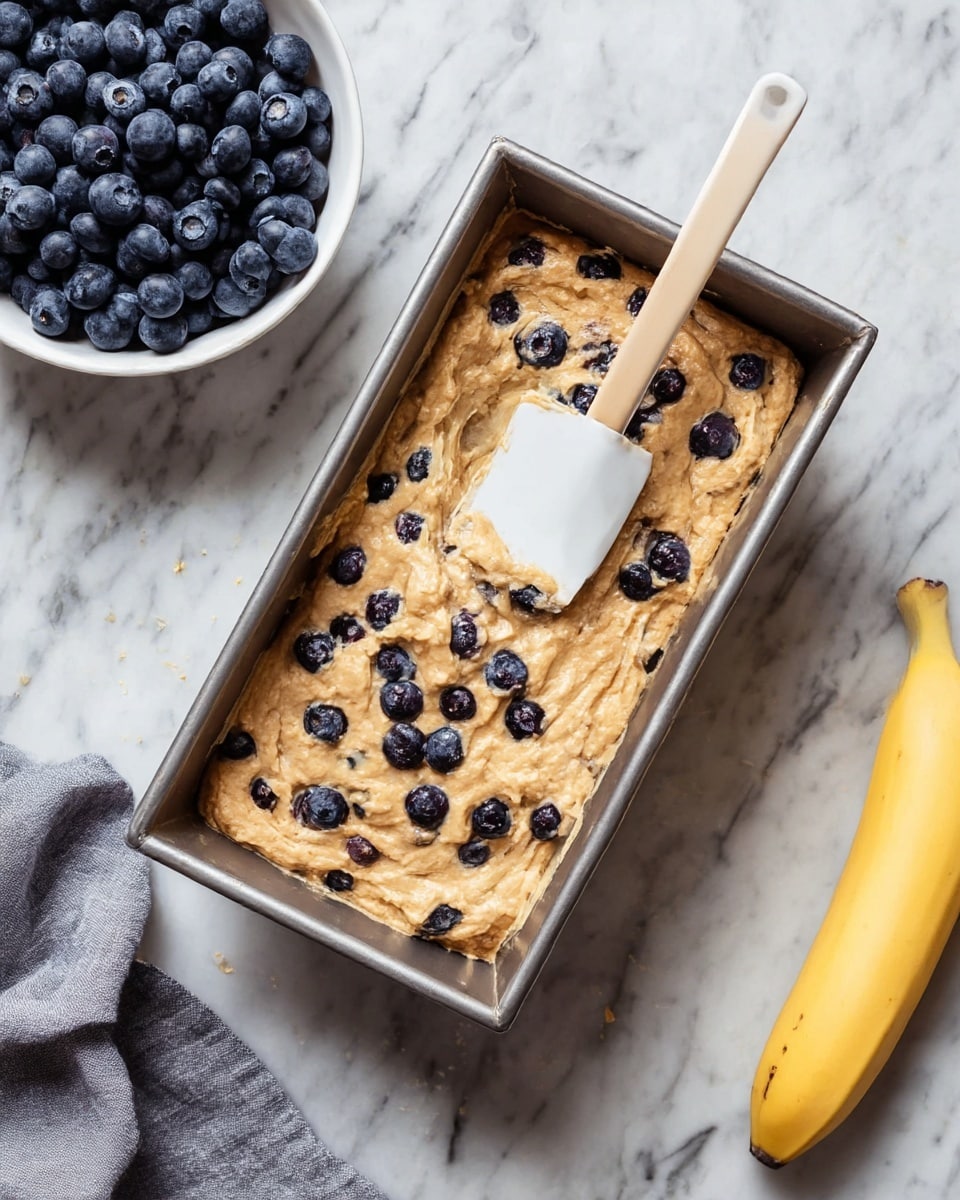 A metal rectangular loaf pan filled with thick, light brown batter mixed with whole blueberries spread unevenly on top. A white spatula rests inside the pan, partially covered with batter, placed diagonally near the center. To the left, there is a white bowl filled with fresh blueberries, and to the right, a yellow banana lies on the white marbled surface. A folded gray cloth is placed near the bottom left corner. photo taken with an iphone --ar 4:5 --v 7