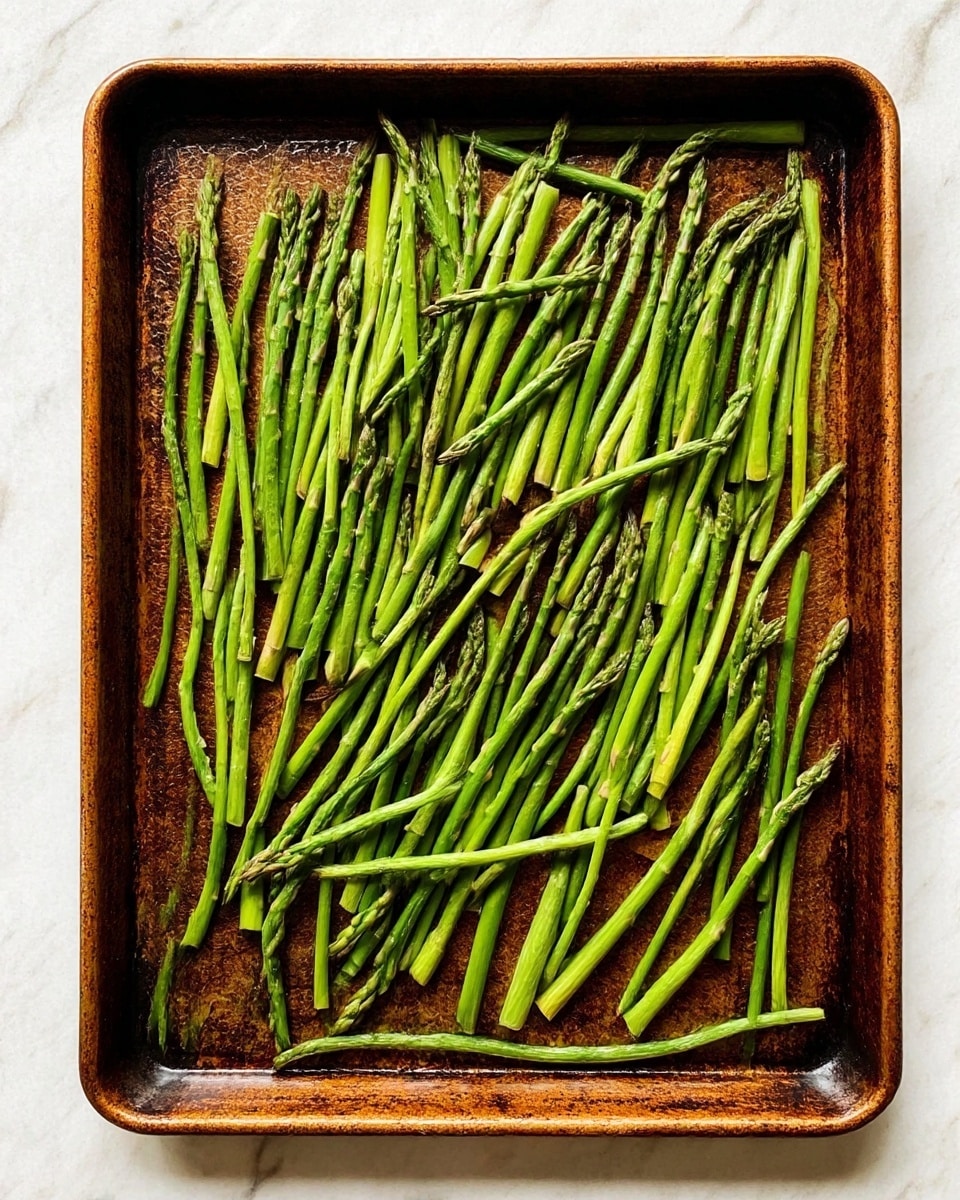 The image shows a rectangular metal baking tray filled with many green asparagus stalks spread in one even layer. The asparagus stalks are thin and long, laid out mostly parallel, with some overlapping slightly. The baking tray has a textured, dark brown surface with signs of use and slight discoloration from roasting. The background is a white marbled surface. photo taken with an iphone --ar 4:5 --v 7