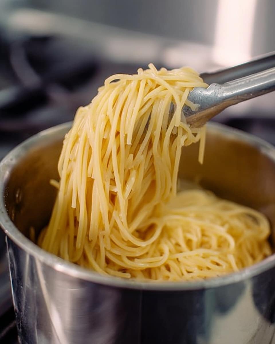A close-up image shows a silver pot filled with plain cooked spaghetti noodles. The noodles are light yellow and smooth in texture, lifted above the pot by silver tongs. The background is blurred and metallic, emphasizing the shiny, open pot on a stove. Photo taken with an iphone --ar 4:5 --v 7