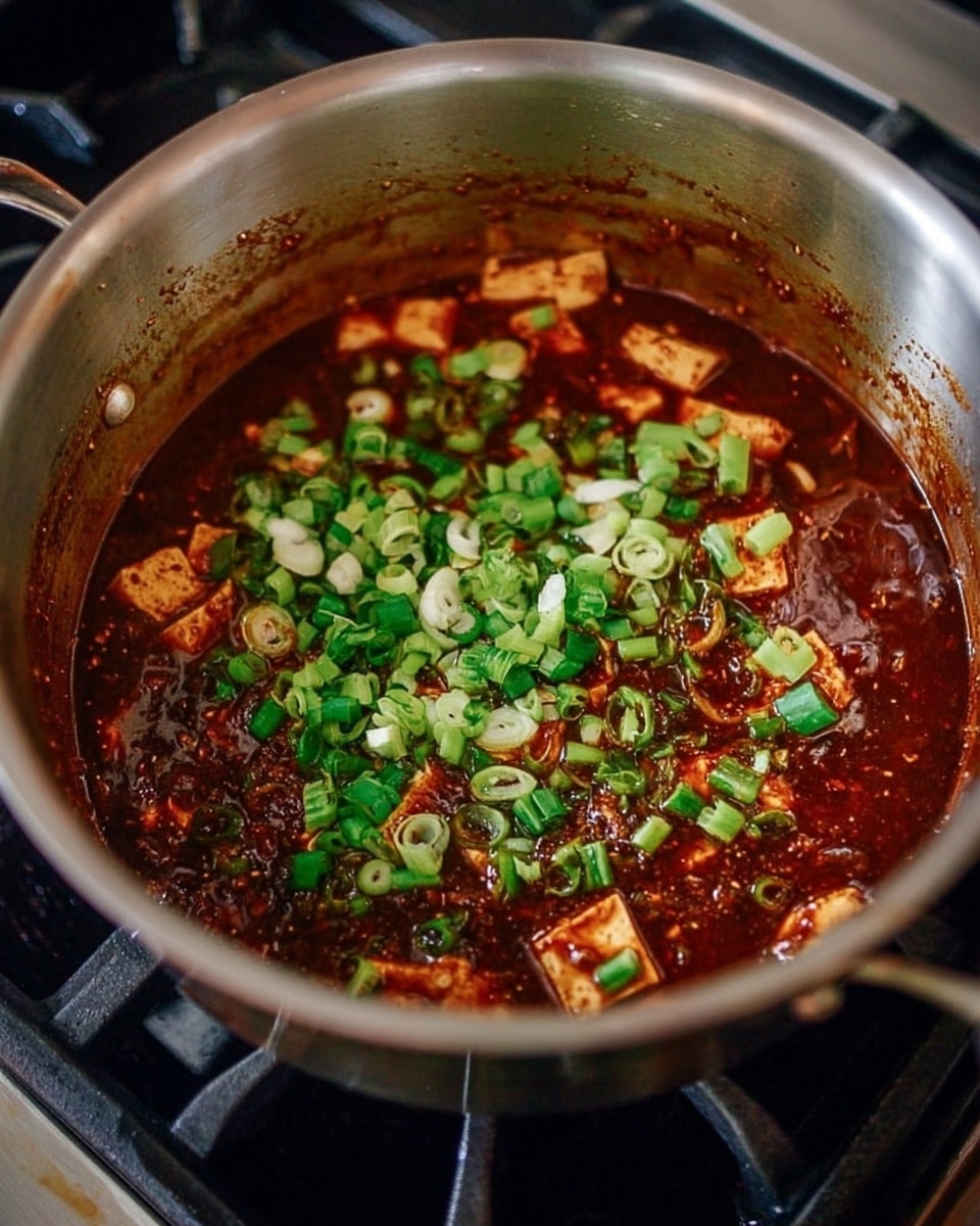 Inside a large stainless steel pan, there is a rich, dark red sauce with a thick texture swirling around pieces of light brown tofu. On top of the sauce and tofu, there are fresh green and white chopped scallion slices spread evenly. The pan sits on a black stovetop, and the inside of the pan shows some sauce splatters on the sides, adding to the cooking scene. Photo taken with an iphone --ar 4:5 --v 7
