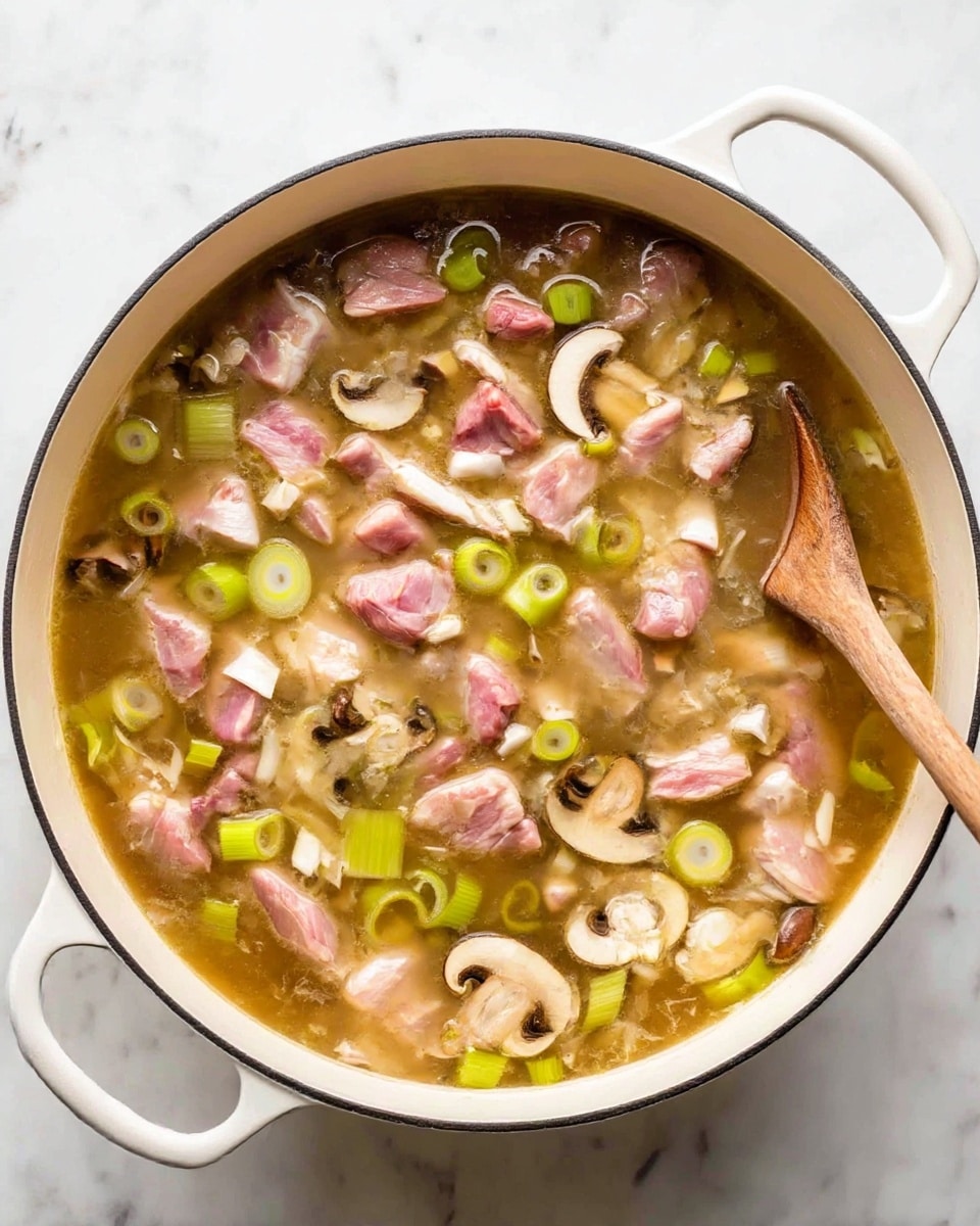 The image shows a large white pot filled with a broth that has a light brown color. Inside the broth, there are many pieces of raw pink meat scattered evenly, with some white fat parts visible. There are also light green circular slices of leek and light brown sliced mushrooms floating in the broth. A wooden spoon is partially submerged in the pot on the right side, stirring the ingredients gently. The pot is placed on a white marbled surface. photo taken with an iphone --ar 4:5 --v 7
