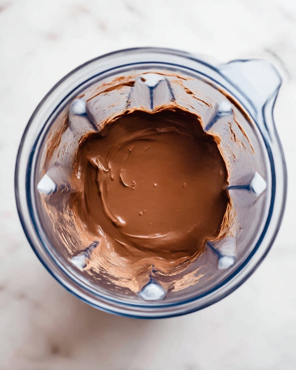 Inside a clear blender jar, there is one smooth layer of thick, creamy chocolate mixture with a rich brown color and a shiny surface. The chocolate sits centrally, spreading evenly to the sides of the blender's sharp inner contours. The container’s plastic walls are clear, showing a bit of chocolate residue near the top edges. The blender is placed on a white marbled surface. photo taken with an iphone --ar 4:5 --v 7