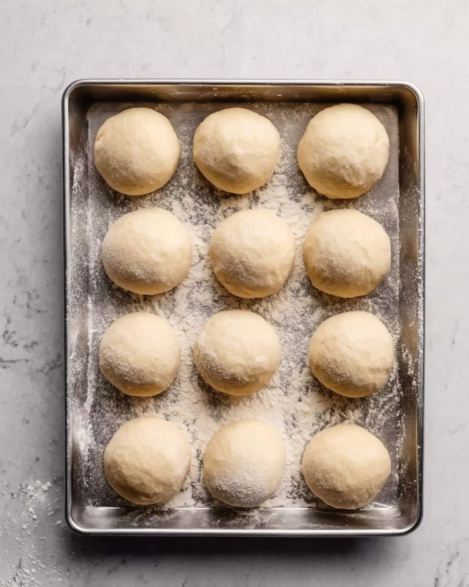 The image shows a metal tray with twelve smooth, round dough balls evenly spaced in three rows and four columns. The dough balls are pale beige with a soft texture and a light dusting of white flour on and around them. The tray rests on a white marbled surface that gives a clean and bright background to the scene. photo taken with an iphone --ar 4:5 --v 7