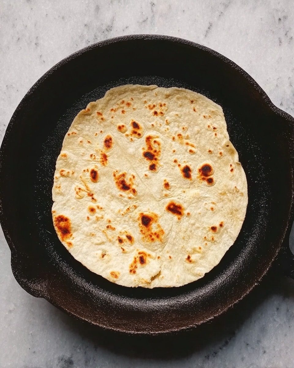 A single flat, round tortilla with light brown spots is cooking in a dark cast iron pan. The tortilla's surface is slightly uneven, with some small bubbles and charred marks, showing a soft texture. The pan has a worn, rustic look with ridged edges. The background is a white marbled surface. photo taken with an iphone --ar 4:5 --v 7