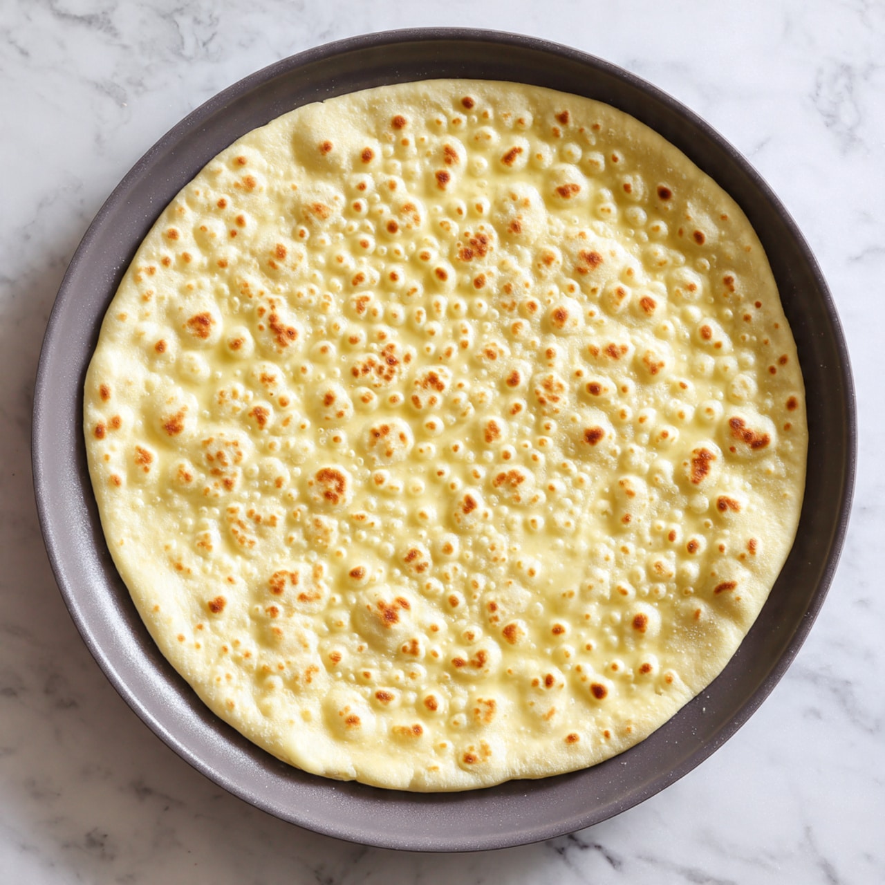 A single round dough flatbread cooking on a smooth dark gray pan with multiple small and large bubbles forming on its surface; the dough is pale yellow with a soft, slightly uneven texture, and the flatbread is centered in the pan against a white marbled surface background photo taken with an iphone --ar 4:5 --v 7
