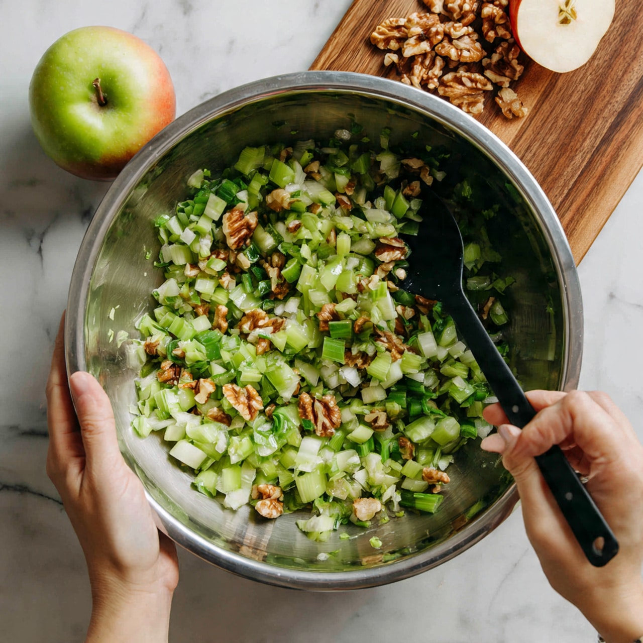 A large stainless steel bowl filled with chopped celery, small pieces of walnuts, and bits of green onions is being mixed. The green celery pieces are the main visible layer, with darker green rings of green onions scattered through. Bits of light and medium brown walnuts add texture and color contrast to the green vegetables. A woman's hand holds a black spoon inside the bowl stirring the mix, while the other woman's hand steadies the bowl. The bowl is placed on a white marbled textured surface, next to a wooden board with half of an apple resting on it. photo taken with an iphone --ar 4:5 --v 7