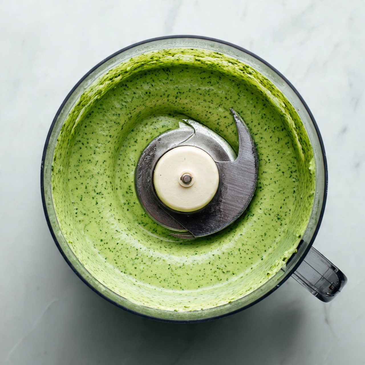 A clear round food processor bowl shows a smooth green sauce with small dark green herb bits mixed evenly throughout. The sauce fills about half the bowl and has a creamy texture, surrounding a curved silver blade with a white plastic center in the middle. The surface beneath is a white marbled texture. photo taken with an iphone --ar 4:5 --v 7