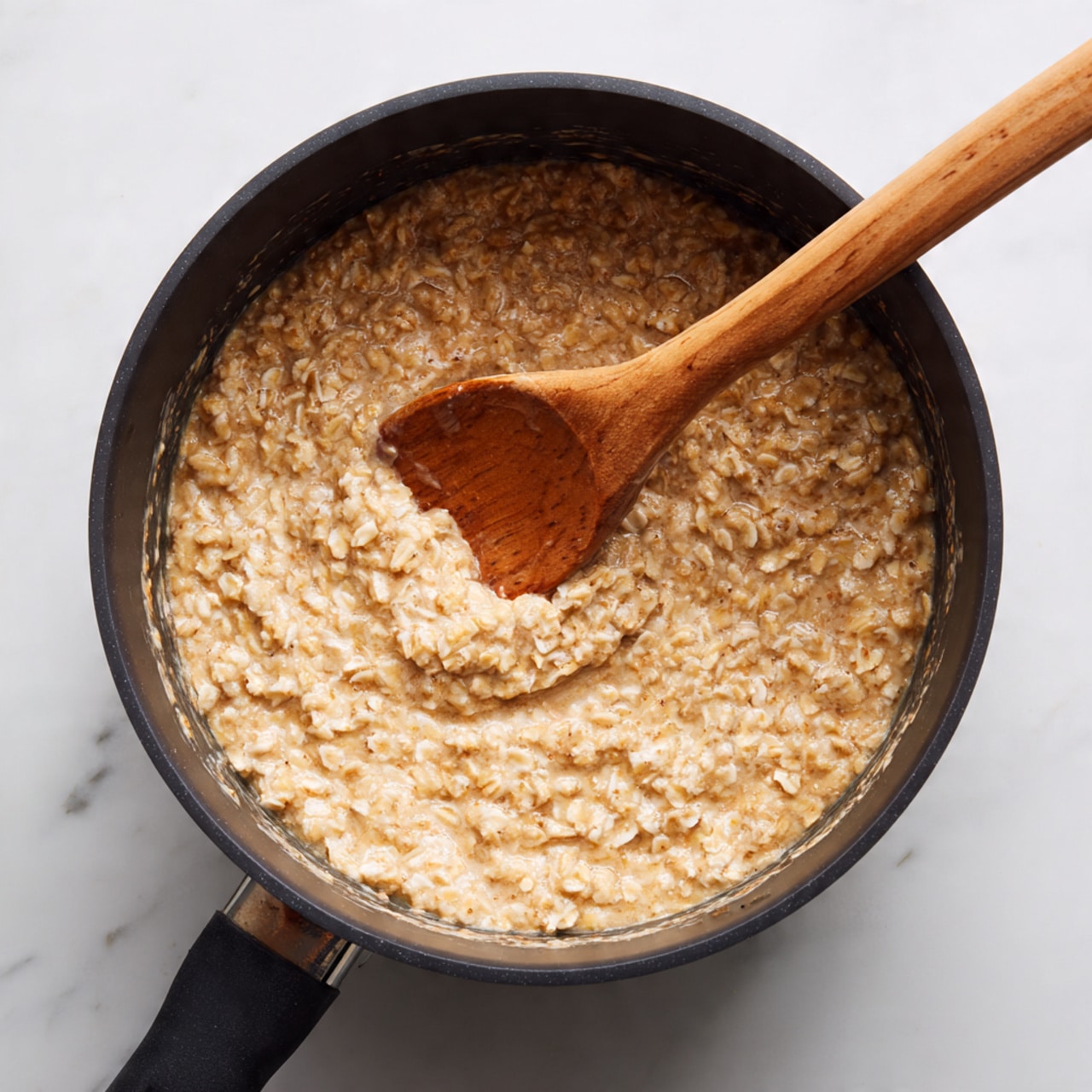 A close-up view of a black pot filled with sticky oatmeal mixture, showing a thick, textured layer of light brown oats evenly spread inside. A wooden spoon is partially submerged in the oatmeal on the right side, its handle resting on the pot’s edge, stirring the oats. The pot is sitting on a white marbled surface. photo taken with an iphone --ar 4:5 --v 7
