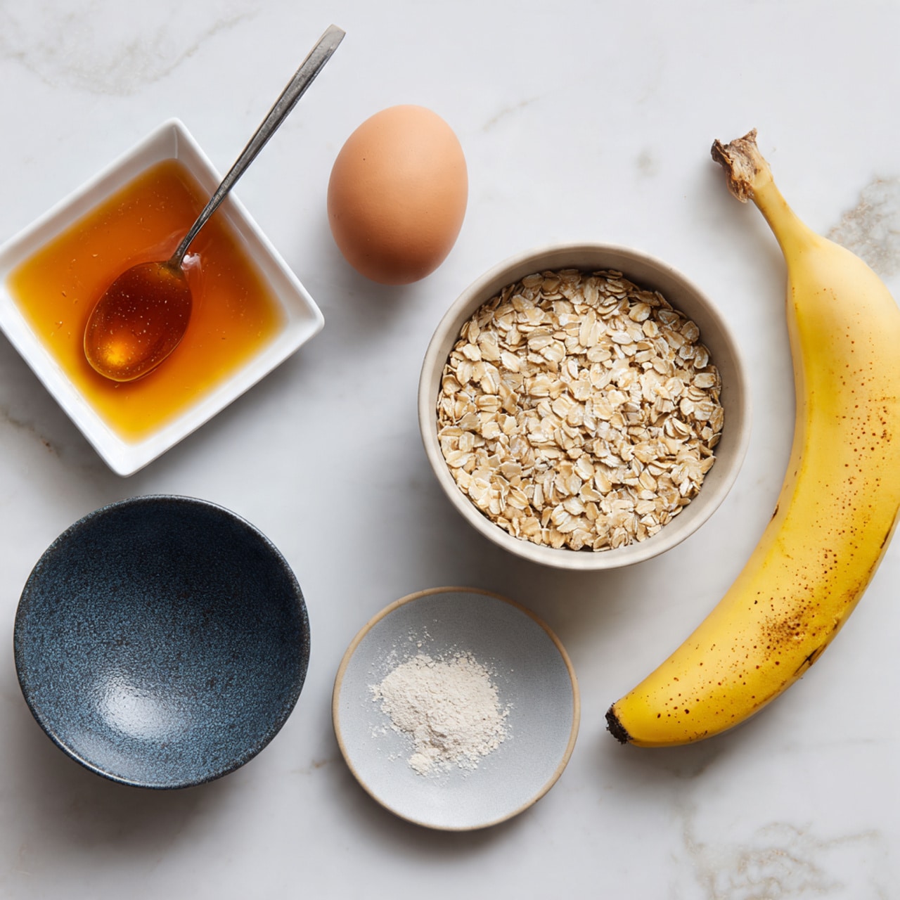 The image shows five ingredients placed on a white marbled surface: a white small square dish filled with amber-colored syrup and a silver spoon resting in it at the top left; below it, a small dark blue speckled bowl holding a single brown egg; in the top center, a round white bowl filled with beige, dry rolled oats; below and slightly right, a small white plate with a gray section containing a small heap of white powder; and on the far right, a whole yellow banana with brown spots lying flat. Photo taken with an iphone --ar 4:5 --v 7