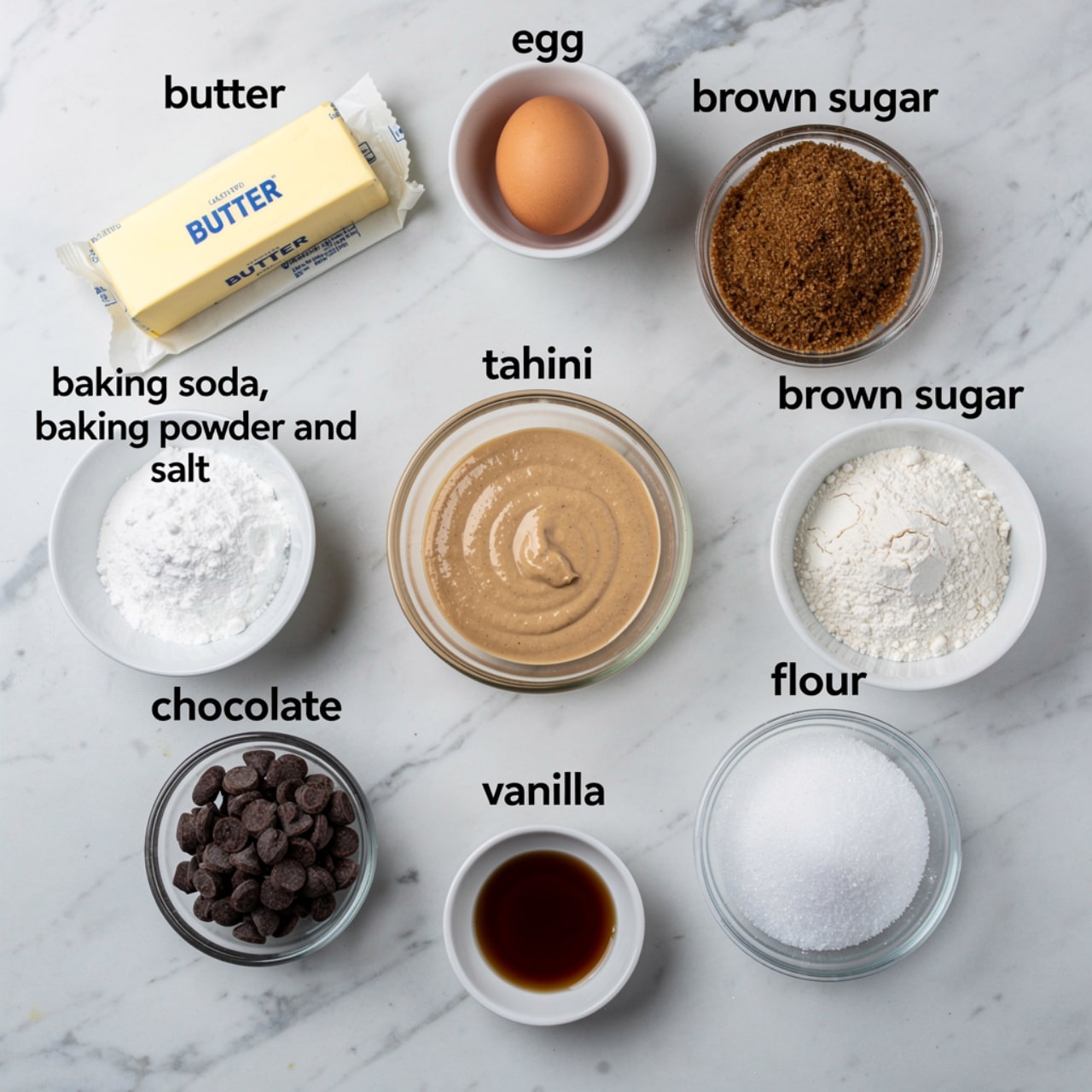 The image shows a recipe layout with nine ingredients placed on a white marbled surface, each in a separate white or clear bowl or plate. At the top center is a small white bowl with one raw egg. To the left, there is a stick of butter in paper wrapping. Below the butter, a small white bowl holds a white powder mix labelled baking soda, baking powder, and salt. In the center is a clear bowl filled with tahini, a smooth light brown paste. To the right of the tahini, a clear bowl contains brown sugar with a coarse texture. Below the brown sugar, a white bowl is filled with loose white flour. Near the bottom right, a clear bowl has granulated white sugar. To the left of that, a small clear bowl contains dark chocolate chips. At the bottom left, a tiny clear bowl holds dark vanilla liquid. Each ingredient is clearly labeled with black text above or beside it. Photo taken with an iphone --ar 4:5 --v 7