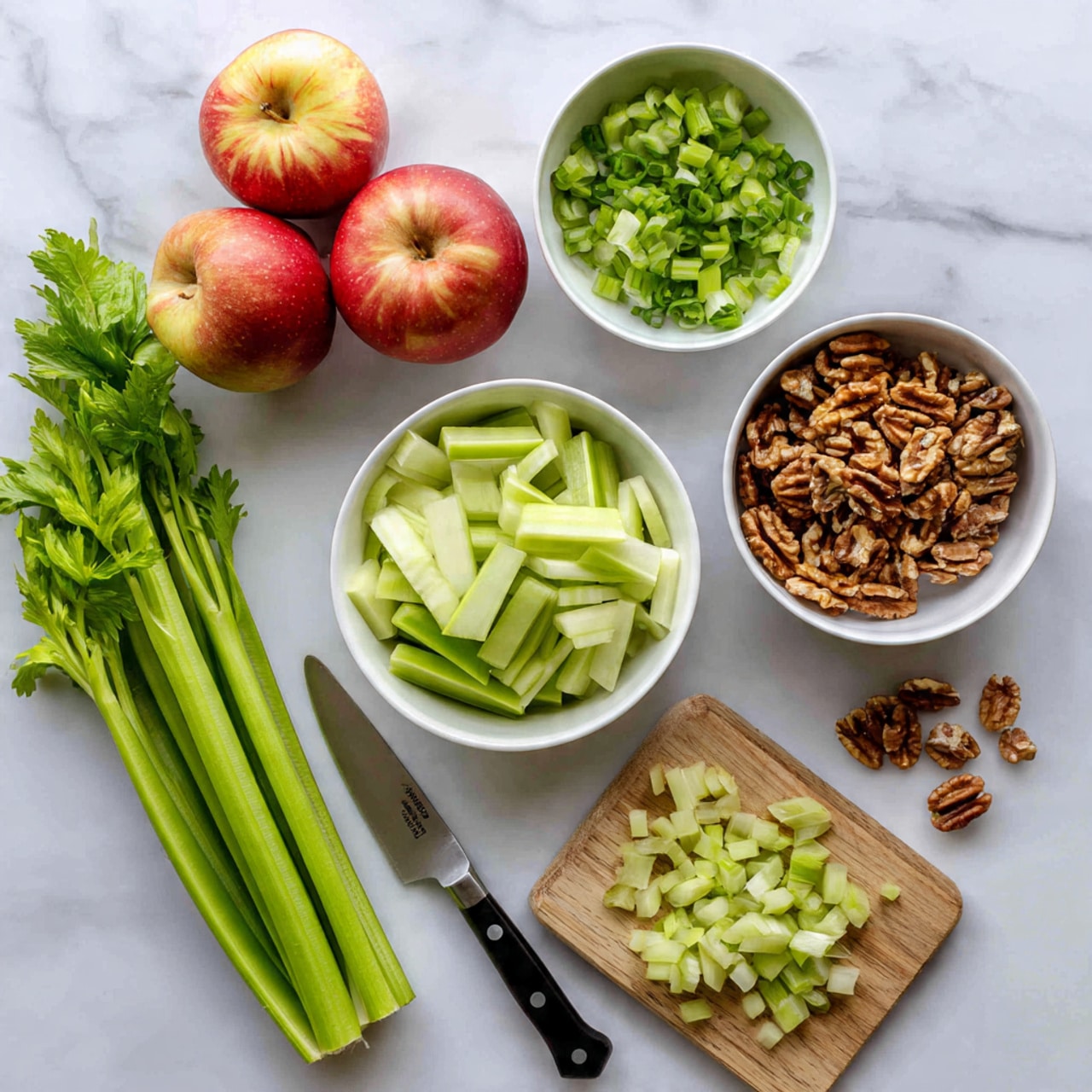 The image shows ingredients arranged on a white marbled surface. At the bottom, there are several fresh green celery stalks with leaves, some chopped celery pieces on a wooden cutting board near a small knife with a black handle. Above the celery, a white bowl is filled with finely chopped celery. To the right of that, there is a white bowl full of small brown walnut pieces, with a few scattered on the surface nearby. Above the walnuts, a white bowl contains thinly sliced bright green scallions. On the upper left side of the image, three pieces of red apple with pale yellow flesh sit directly on the surface. The overall color palette is fresh greens and natural browns with hints of red and pale yellow, all set against the textured white marbled surface. Photo taken with an iphone --ar 4:5 --v 7