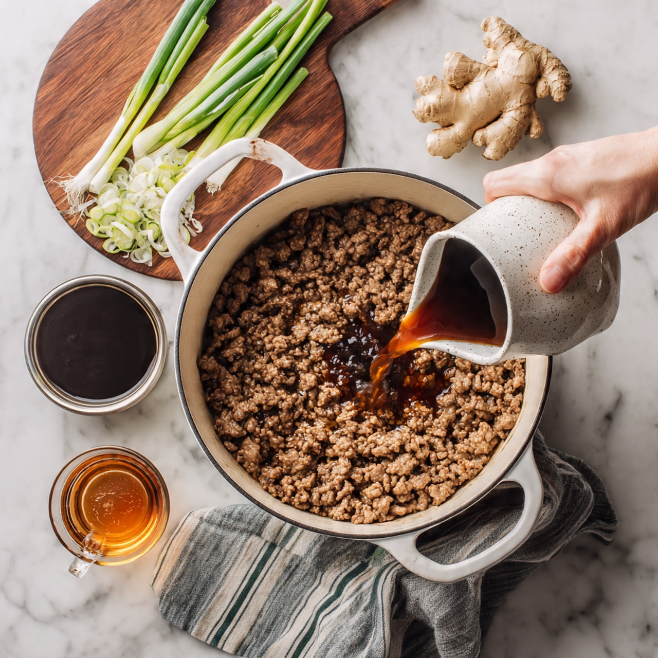 A white pot filled with cooked ground beef, showing a mix of brown and light brown textured meat pieces spread evenly across the pot. A dark brown liquid sauce is being poured from a white speckled jug held by a woman's hand over the meat, creating a shiny wet layer on top. In the back on the white marbled surface, there is a round wooden board with light green whole scallions and white sliced scallions, along with a small bowl filled with thick dark brown sauce. Around the pot are slices of ginger and a glass of amber-colored liquid, with a striped cloth under the pot's handle. Photo taken with an iphone --ar 4:5 --v 7