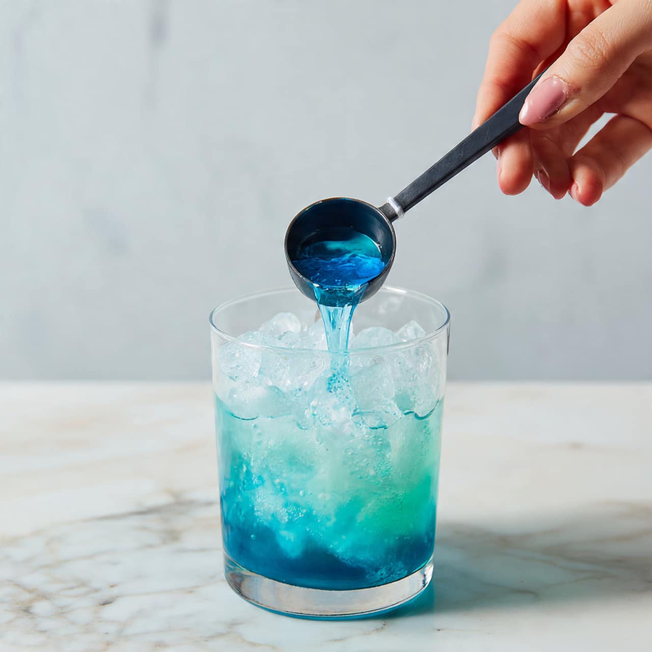 A clear glass filled halfway with ice cubes shows a woman’s hand holding a black measuring spoon above it, pouring a bright blue liquid that cascades into the glass, blending softly with the ice and clear water inside. The blue liquid inside the spoon has a shiny, smooth texture that reflects light, creating a sense of freshness and coolness. The background features a white marbled surface, adding a clean and elegant feel to the image. photo taken with an iphone --ar 4:5 --v 7