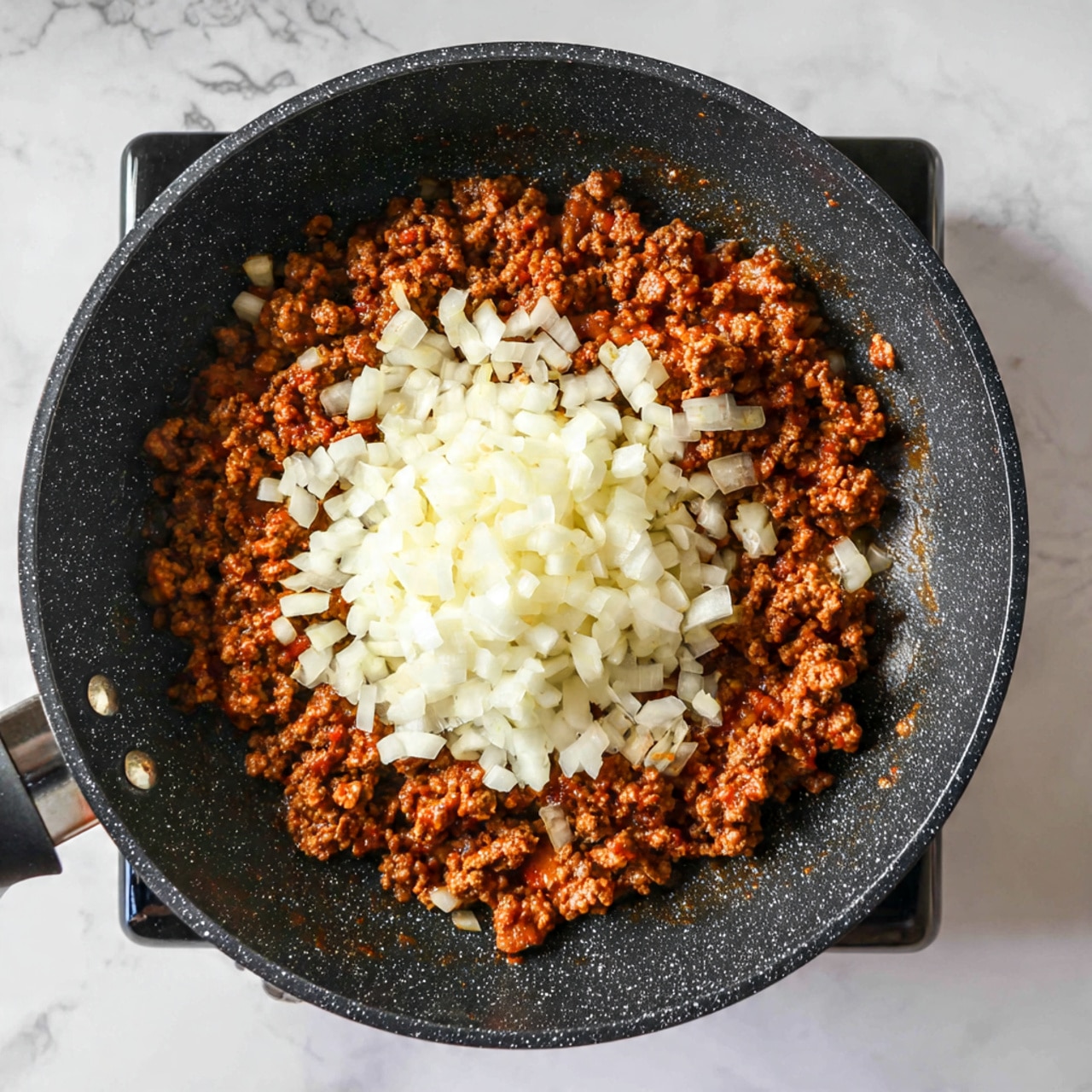 In a large black frying pan with a speckled non-stick surface, there is one layer of browned ground meat spread evenly across the pan. On top, in the center, there is a single layer of chopped white onions with a crisp, slightly translucent texture. The pan is sitting on a stove with a white marbled surface visible around it. photo taken with an iphone --ar 4:5 --v 7