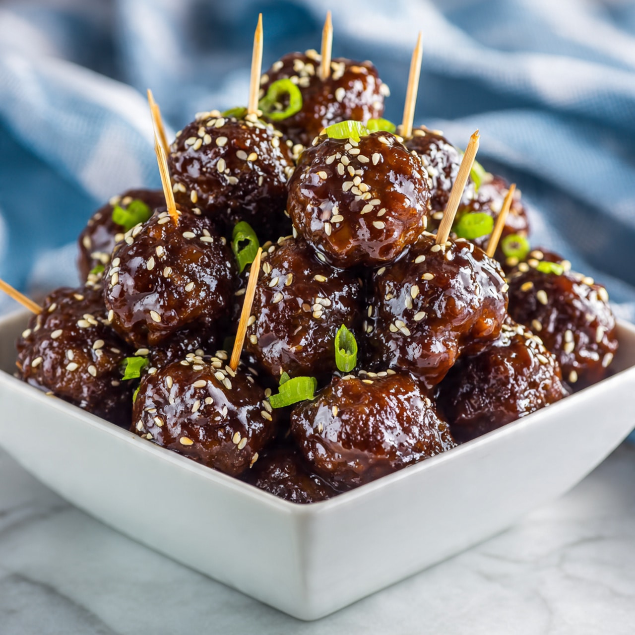 A white square bowl is filled with many glossy dark brown meatballs covered in a sticky sauce. The meatballs are stacked high and topped with light tan sesame seeds and small rings of green onions scattered across. Several toothpicks stick out of the meatballs, showing they are ready for picking. The bowl sits on a white marbled texture with a blurred blue and white striped cloth in the background. photo taken with an iphone --ar 4:5 --v 7
