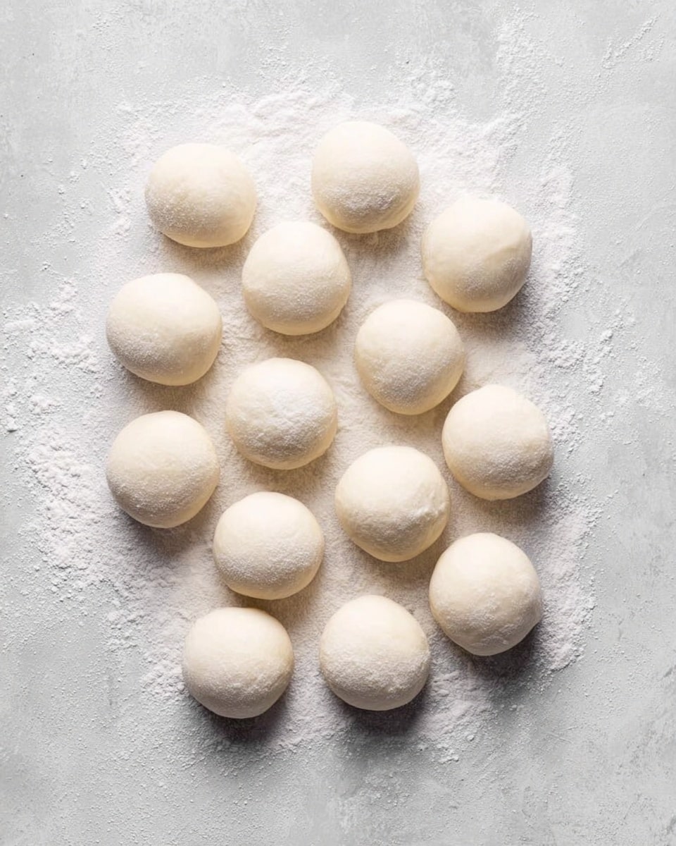 The image shows fourteen small, round dough balls placed directly on a light gray surface with some white flour dusted underneath and around them. Each dough ball is smooth and white, evenly spaced with soft shadows beneath, giving a fresh and soft texture look. The background has a slightly rough but clean texture that complements the dough balls. photo taken with an iphone --ar 4:5 --v 7