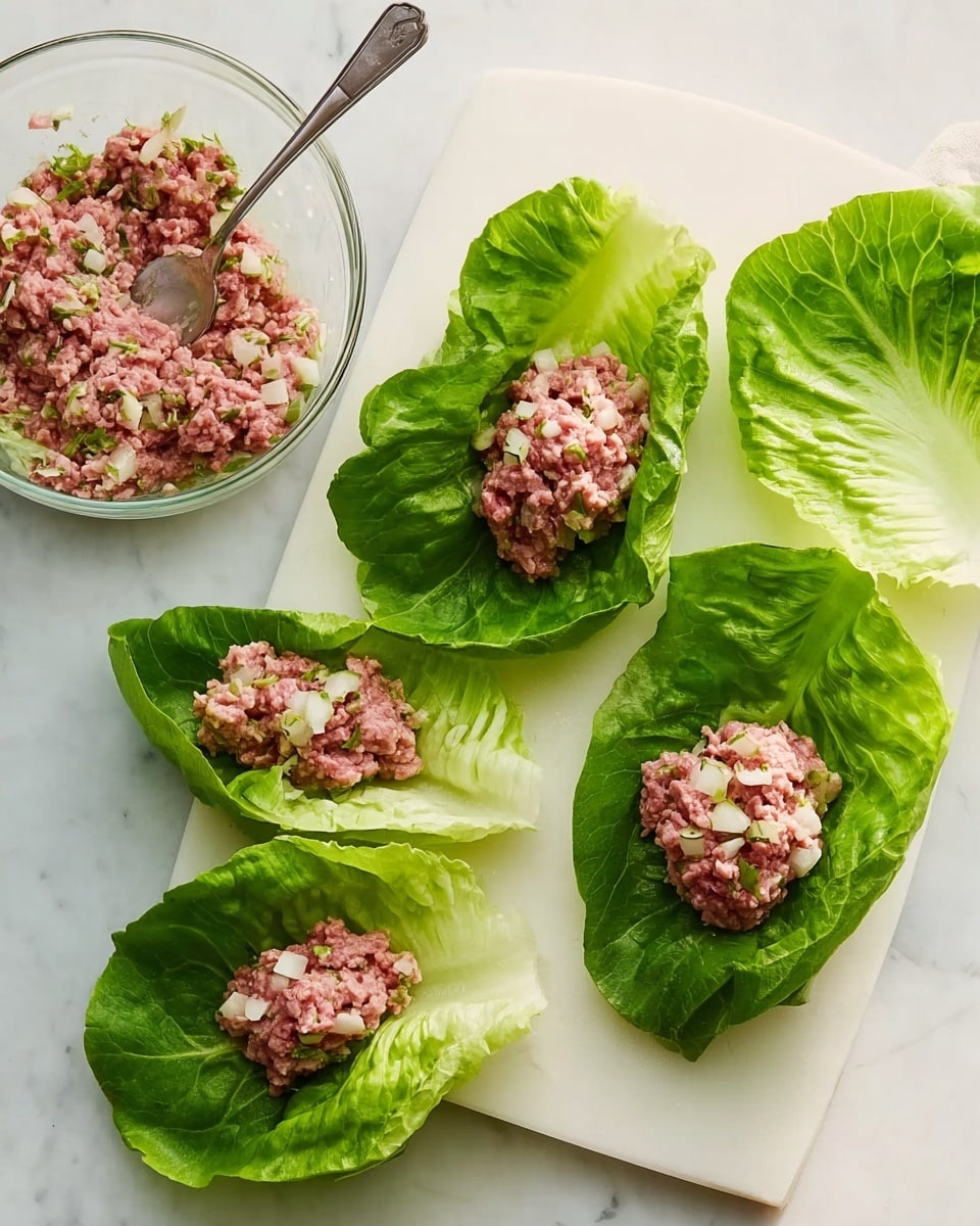 The image shows a white cutting board on a white marbled surface, with four large green lettuce leaves neatly placed on it. Three of these leaves are filled with a mix of pinkish raw ground meat and small white bits, positioned in the center of each leaf, while the fourth leaf is empty. In the top left corner, there is a transparent glass bowl containing more of the meat mixture with a silver spoon resting inside. The overall scene is bright, fresh, and prepared for making a dish, photo taken with an iphone --ar 4:5 --v 7