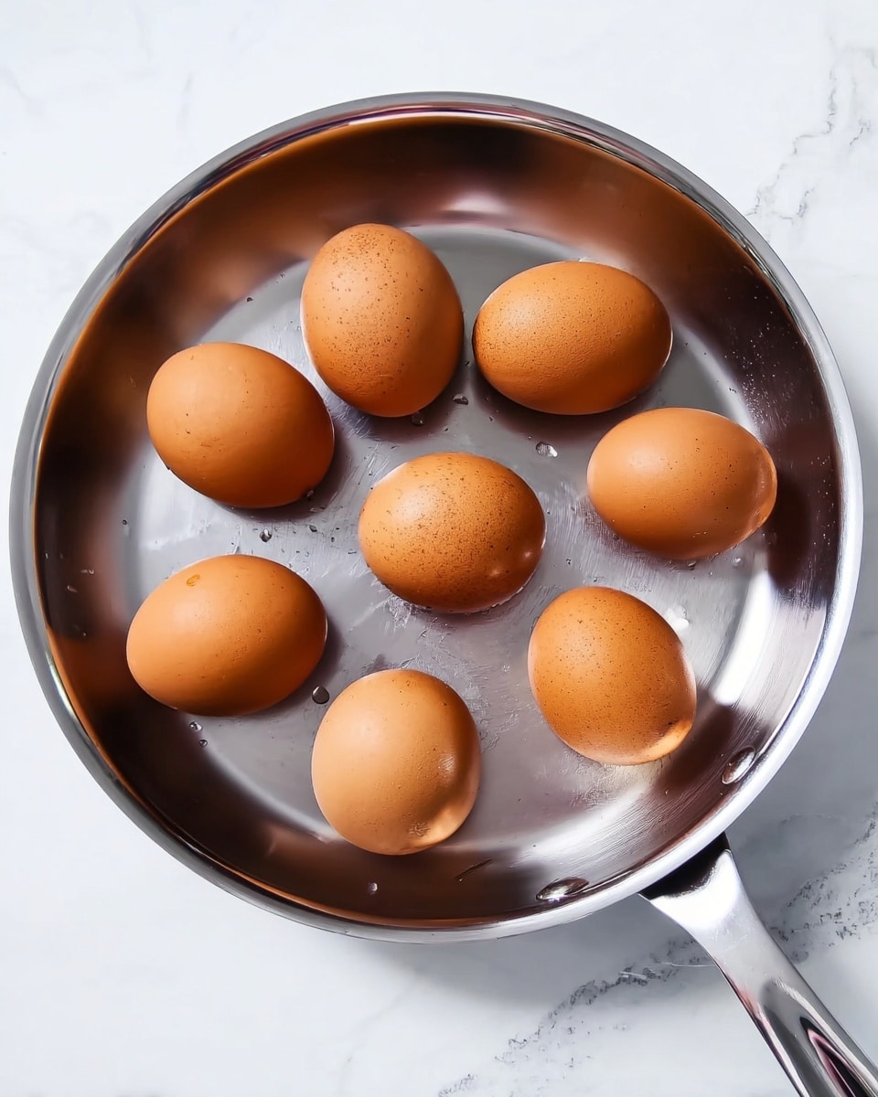A top view of a shiny silver frying pan containing seven whole brown eggs evenly placed inside, on a white marbled surface. The eggs have smooth textures and warm brown colors that contrast with the cool metallic shine of the pan. One egg has a spot of water reflecting light. photo taken with an iphone --ar 4:5 --v 7
