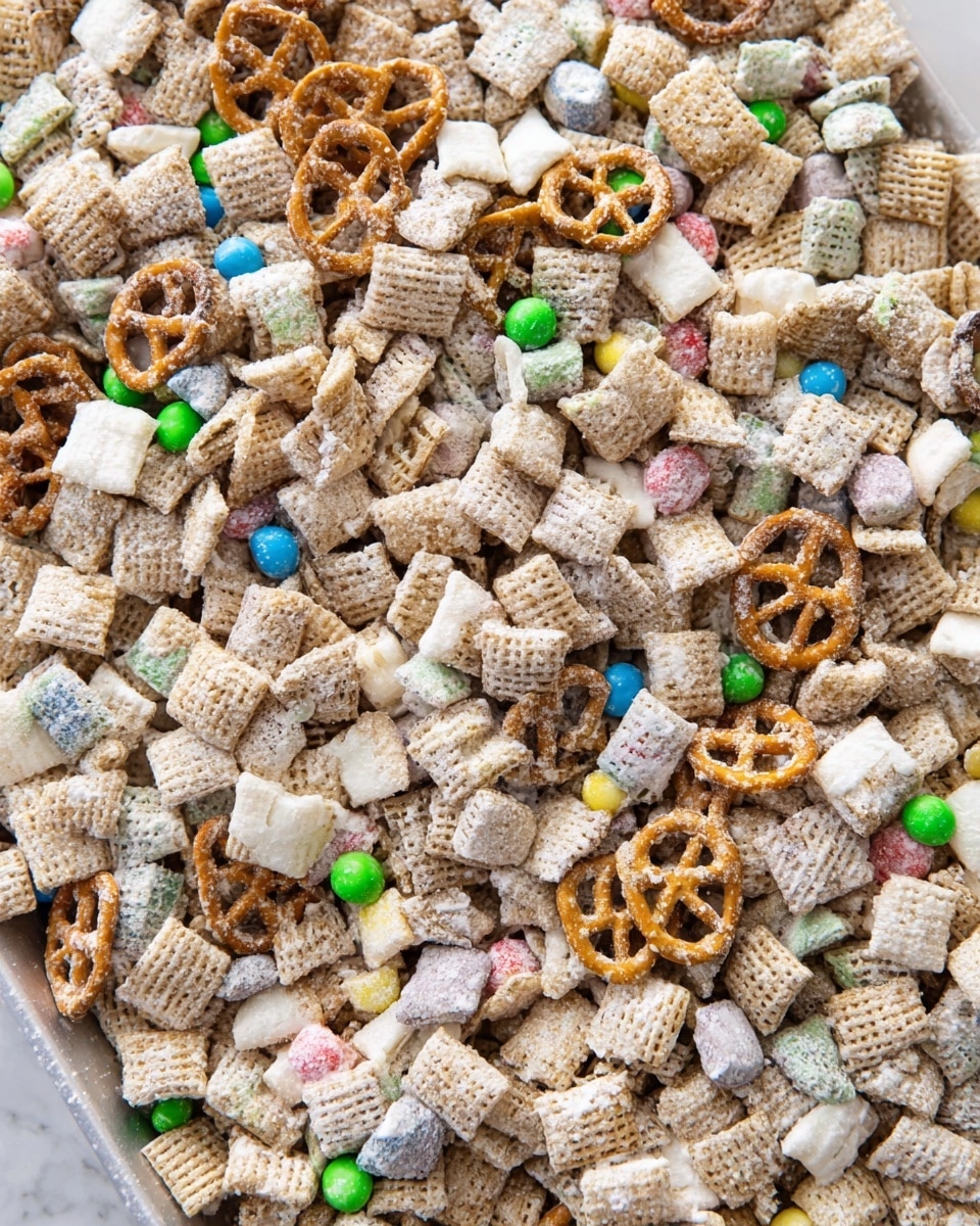 A close-up top view of a white tray filled with a dry crunchy snack mix made of small square cereal pieces, pretzel pieces, and colorful small round candy pieces scattered throughout. The snack mix is coated lightly with a white powdery layer, giving a slightly powdery texture overall. The white marbled surface is visible around the edges of the tray. photo taken with an iphone --ar 4:5 --v 7